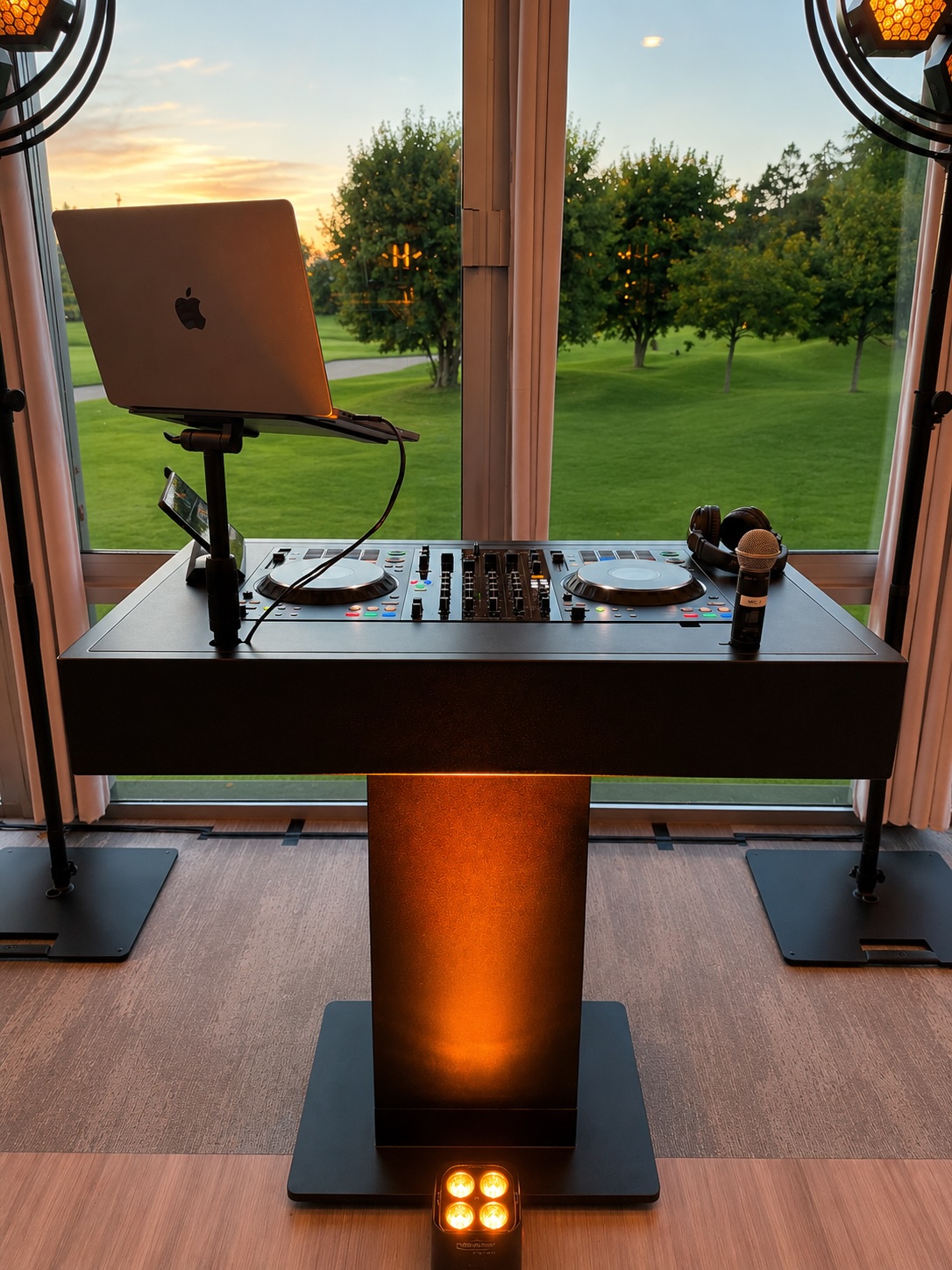 Cabine DJ sur mesure avec pupitre lumineux, face à une baie vitrée donnant sur un parc au coucher du soleil.