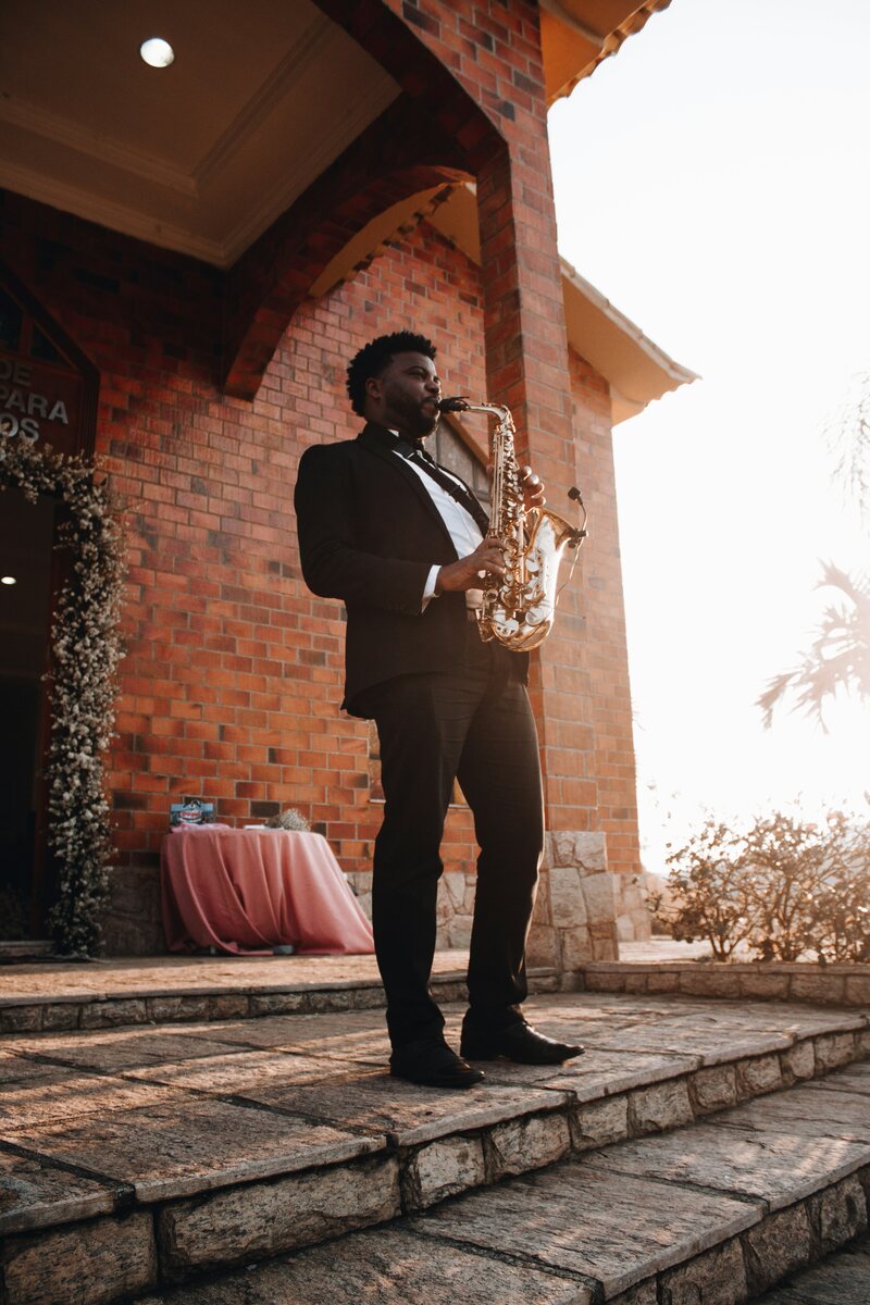 Saxophoniste en costume jouant en extérieur devant un bâtiment en briques, lumière dorée de fin de journée, performance live lors d'un mariage.